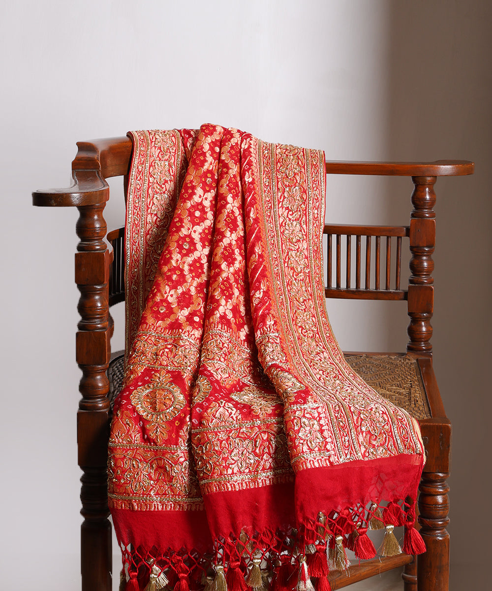 Red bridal Bandhani dupatta draped over a wooden chair, showcasing Banarasi cutwork, zardozi highlights and handcrafted tassels.