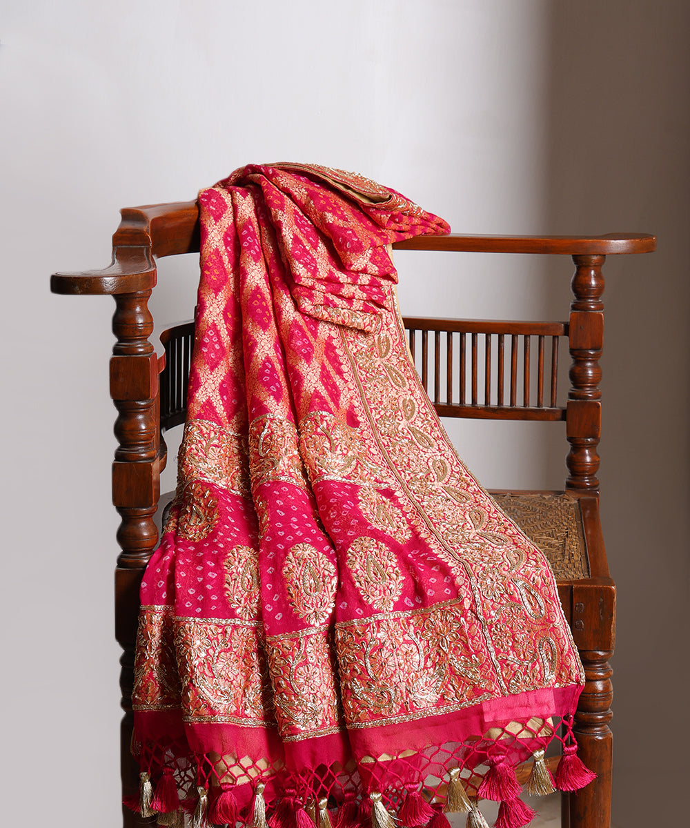 Hot pink bridal Bandhani dupatta draped over a wooden chair, showcasing Banarasi cutwork, rich zardozi borders and handcrafted tassels.