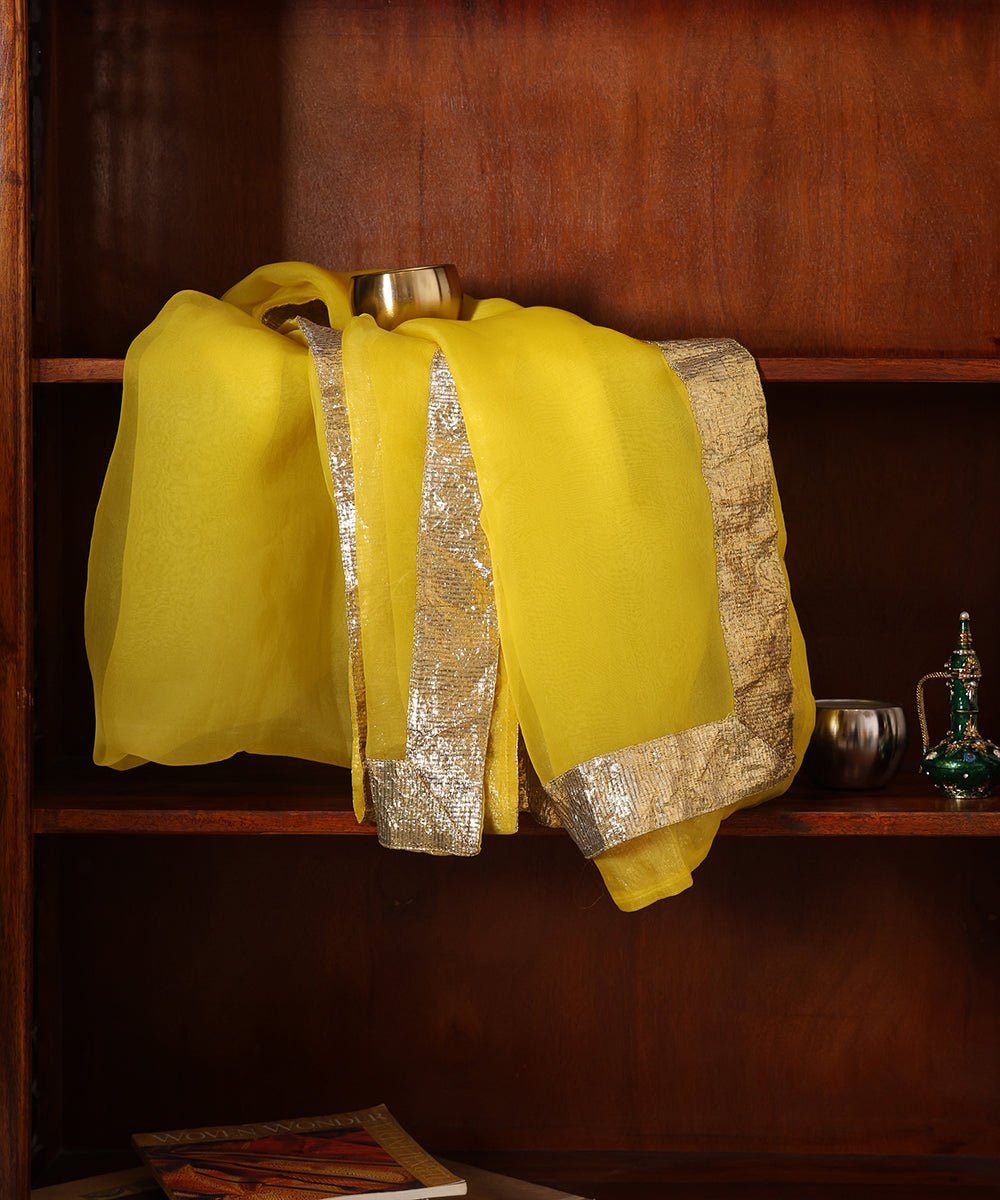 Yellow silk organza dupatta displayed against a wooden cabinet, showing its sheer body and luminous gold lampi gota border