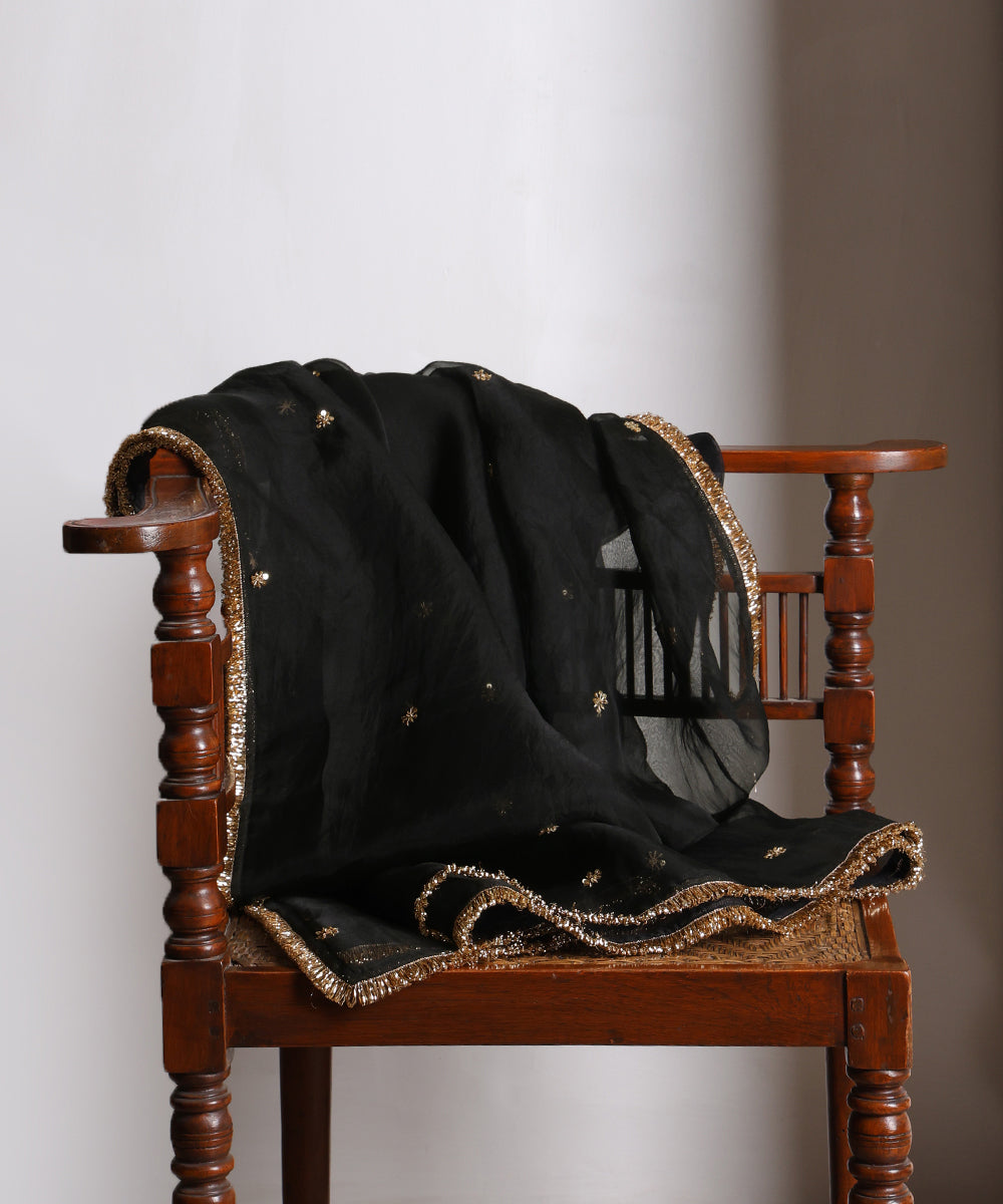 Black organza zardozi dupatta draped over a wooden chair, showing its translucent texture, floral bootis and metallic fringe border.