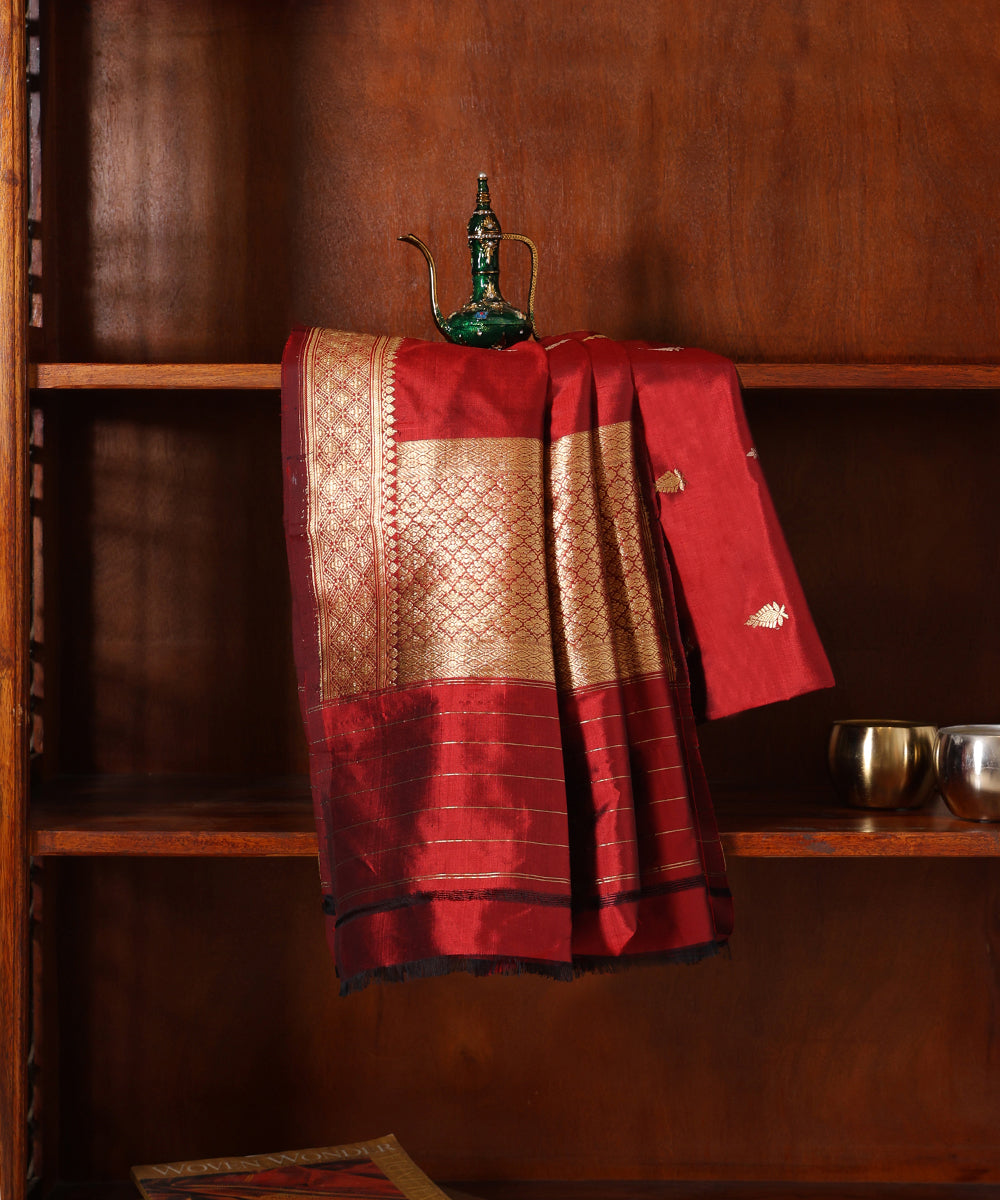 Maroon handloom Katan silk Kadhwa dupatta displayed against a wooden cabinet, featuring zari bootis, a broad geometric palla and striped ends.