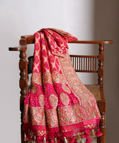 Hot pink bridal Bandhani dupatta draped over a wooden chair, showcasing Banarasi cutwork, rich zardozi borders and handcrafted tassels.