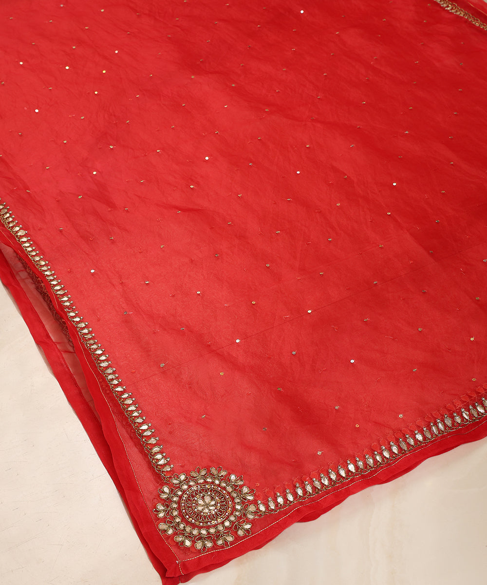 Red silk organza dupatta draped over a wooden chair, showing its sheer texture, gota patti border, scattered sequins and ornate corner embroidery.